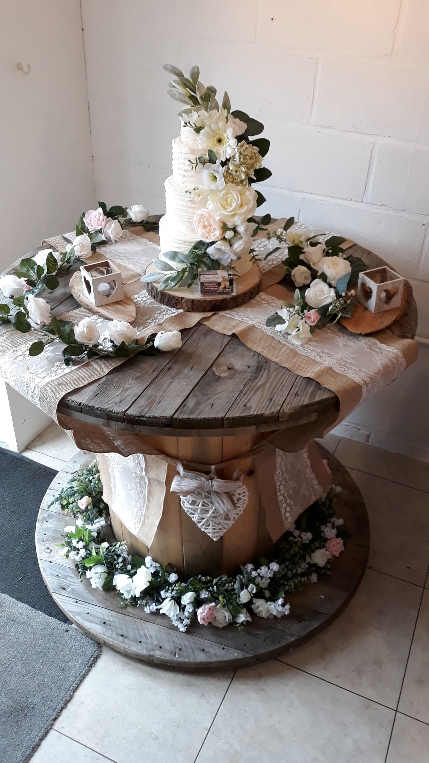 Wedding cake on a rustic wooden spool table, decorated with flowers and lace.
