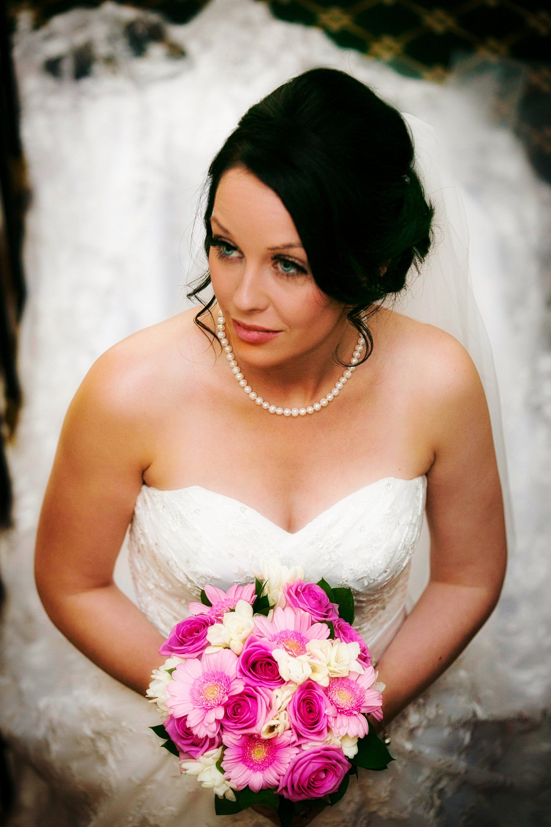 Bride in white strapless dress holding pink and white bouquet, wearing a pearl necklace and veil.