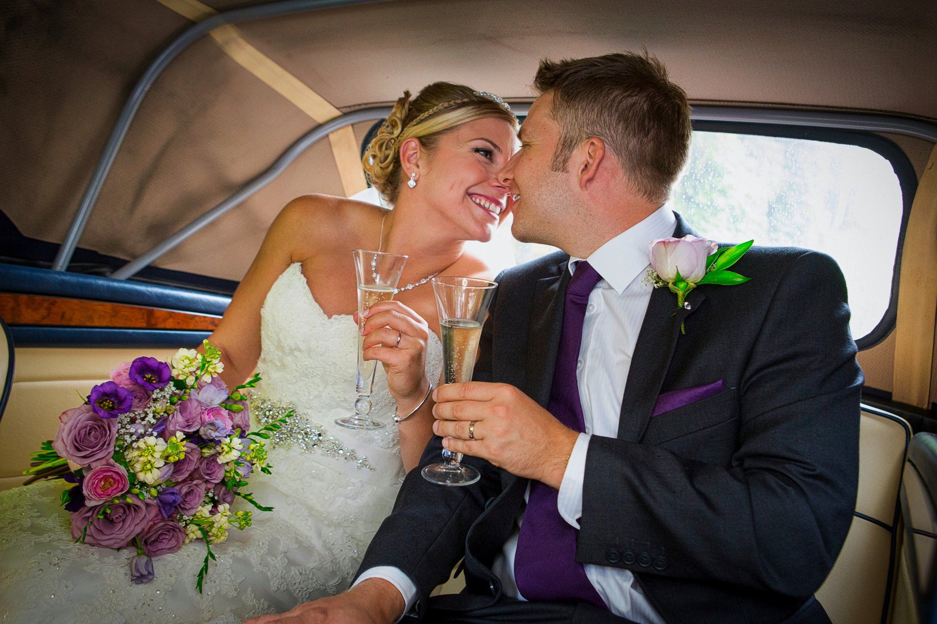 Bride and groom in wedding car, toasting with champagne. The bride smiles, the groom kisses her. Purple flowers.