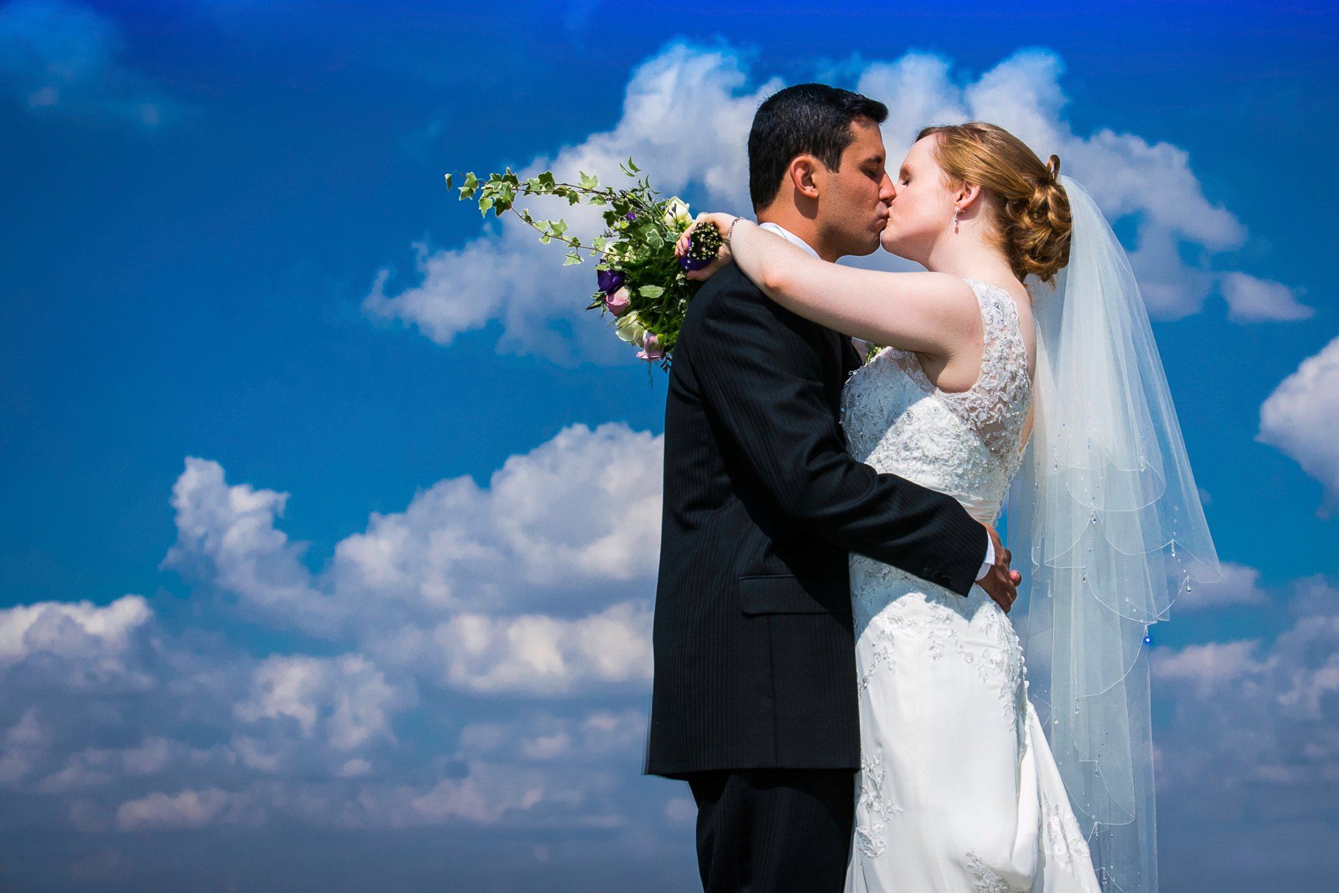 Couple kissing, bride in white dress and veil, groom in black suit, against cloudy blue sky.