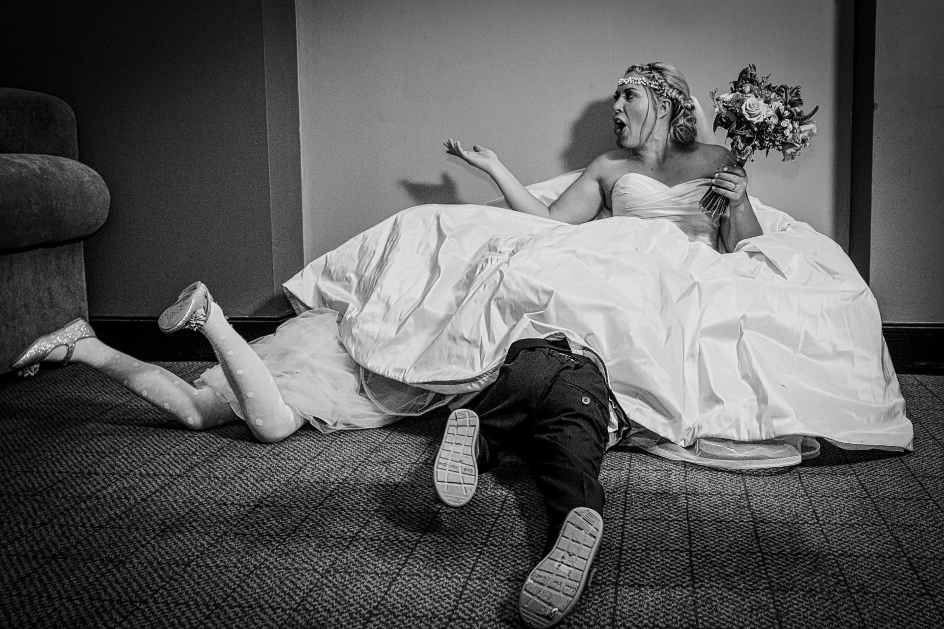 Bride sprawls on floor, dress billowing. Two children, one crawling under the dress, the other lying nearby. Bride looks shocked.