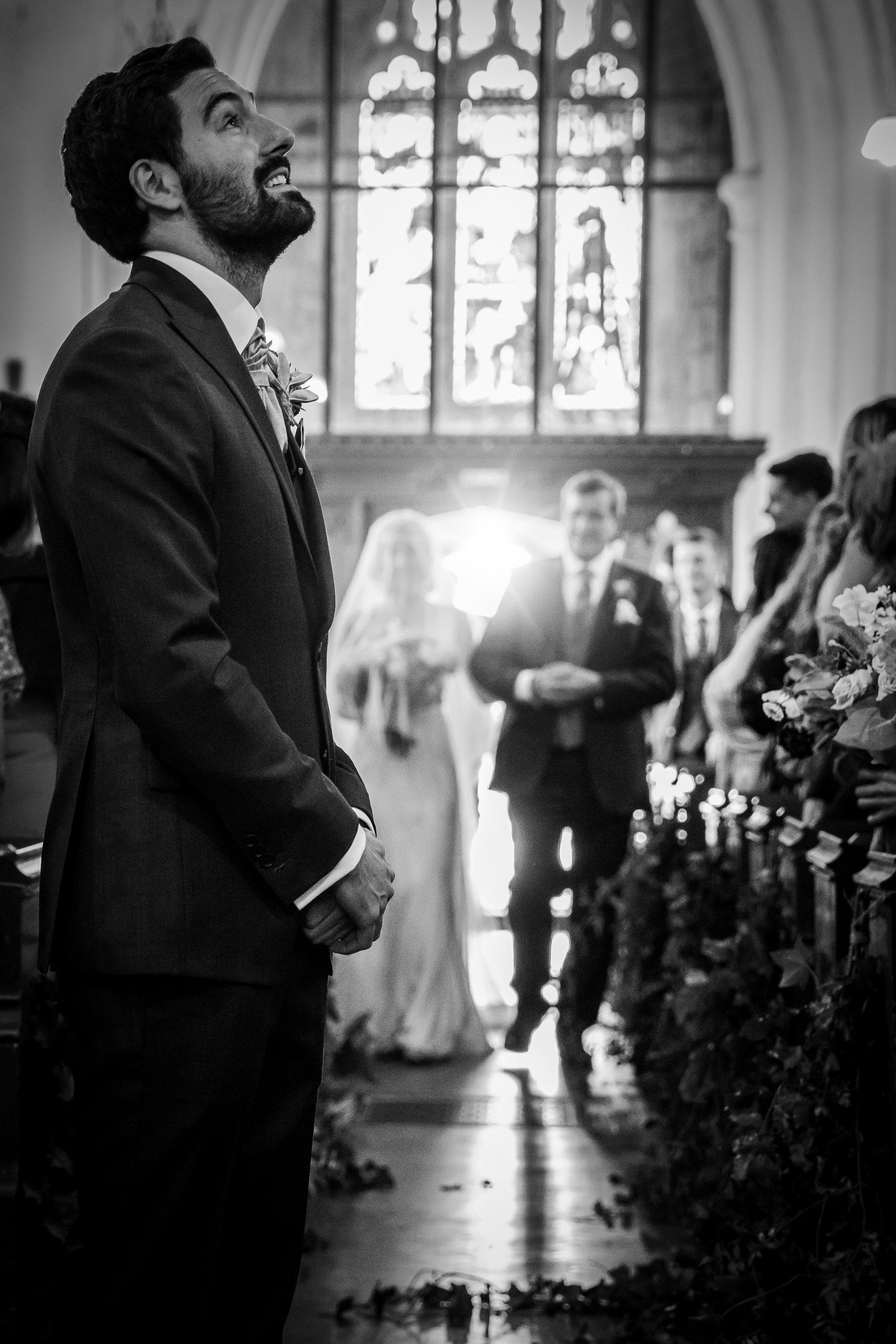 Groom in suit looks up with anticipation as bride walks down the aisle. Church setting, black and white.