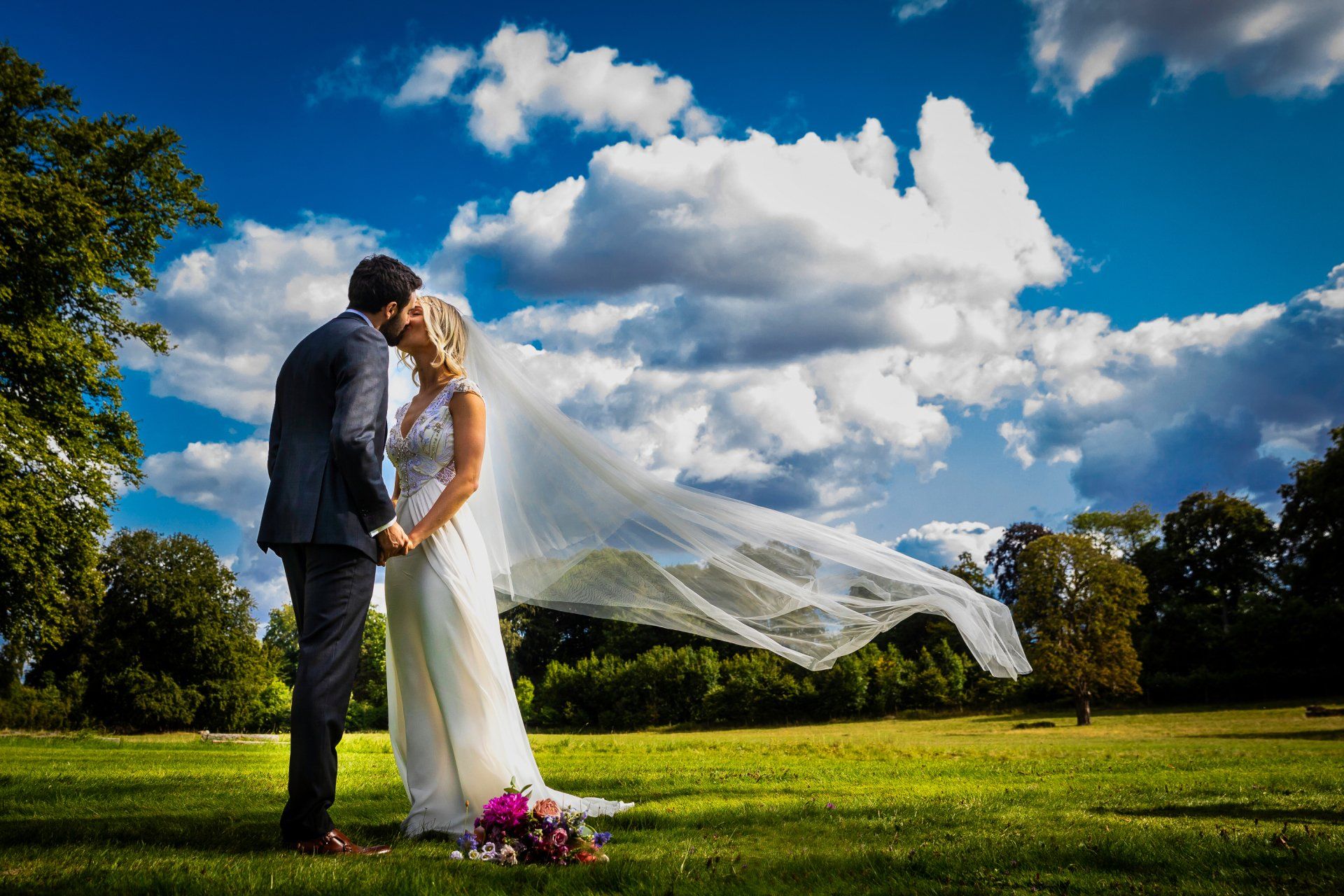 Couple kissing in a grassy field, bride's veil blowing in the wind under a bright blue sky.