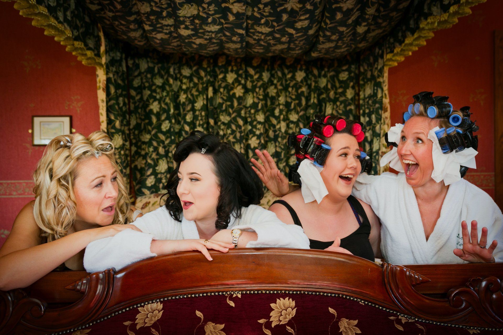 Four women in robes on a bed, laughing, with curlers in their hair. Red walls, patterned curtains.
