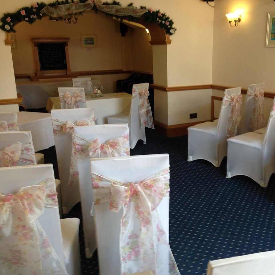 A wedding ceremony setup: white chairs with floral bows, arranged in rows. Ceremony backdrop, neutral colors.