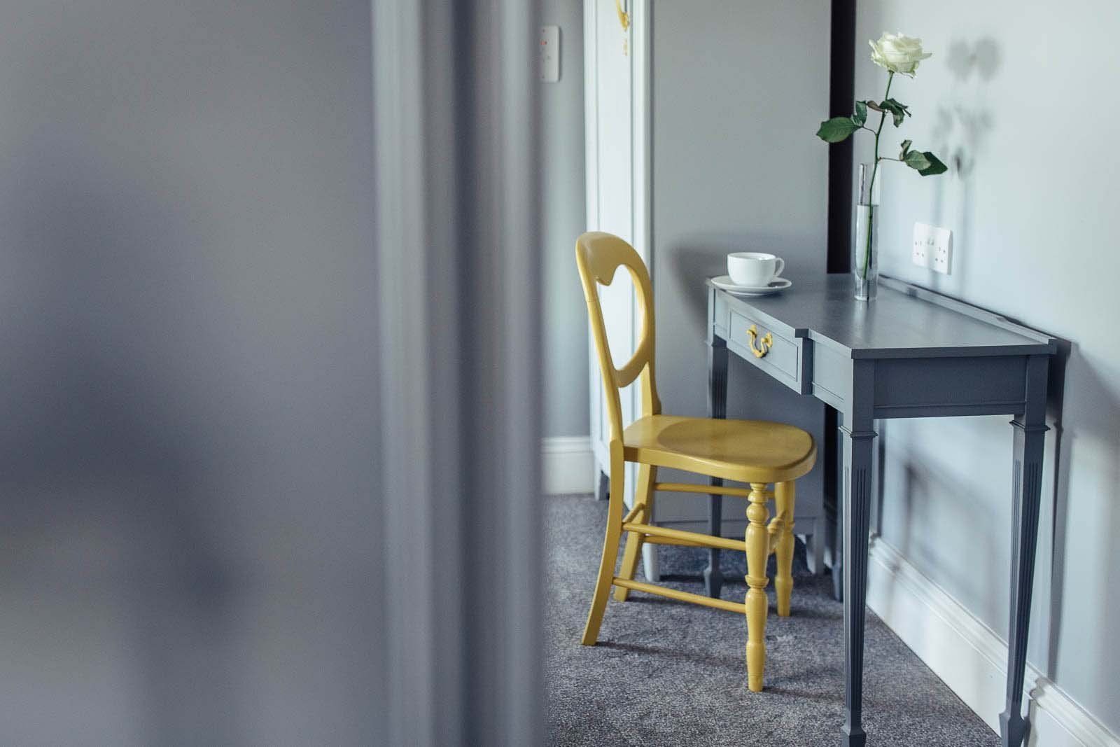 Yellow chair at a desk with a white rose in a vase; light gray walls and carpet.