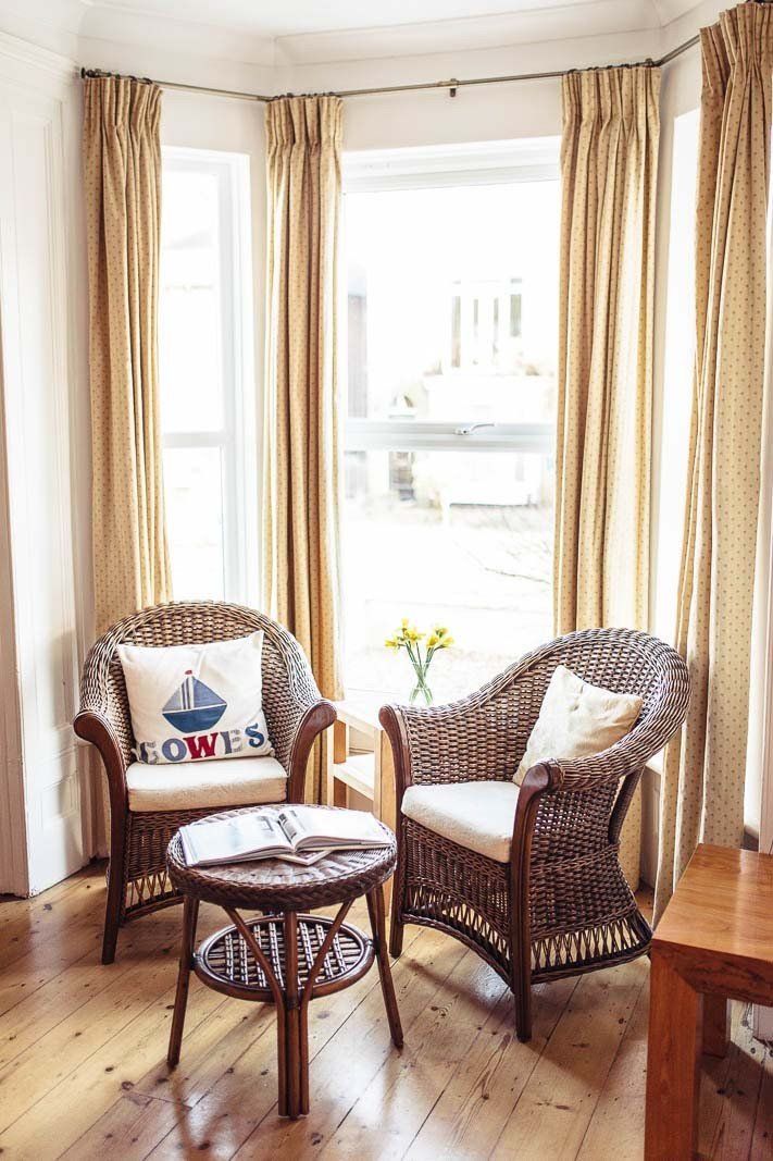 Two wicker chairs, table by a sunny window, beige curtains, nautical pillow.