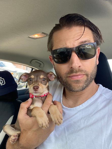 Man in sunglasses holds a small brown and tan dog in a car.
