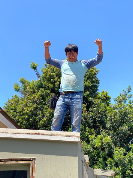 Man on rooftop with arms raised, blue sky, green tree background.