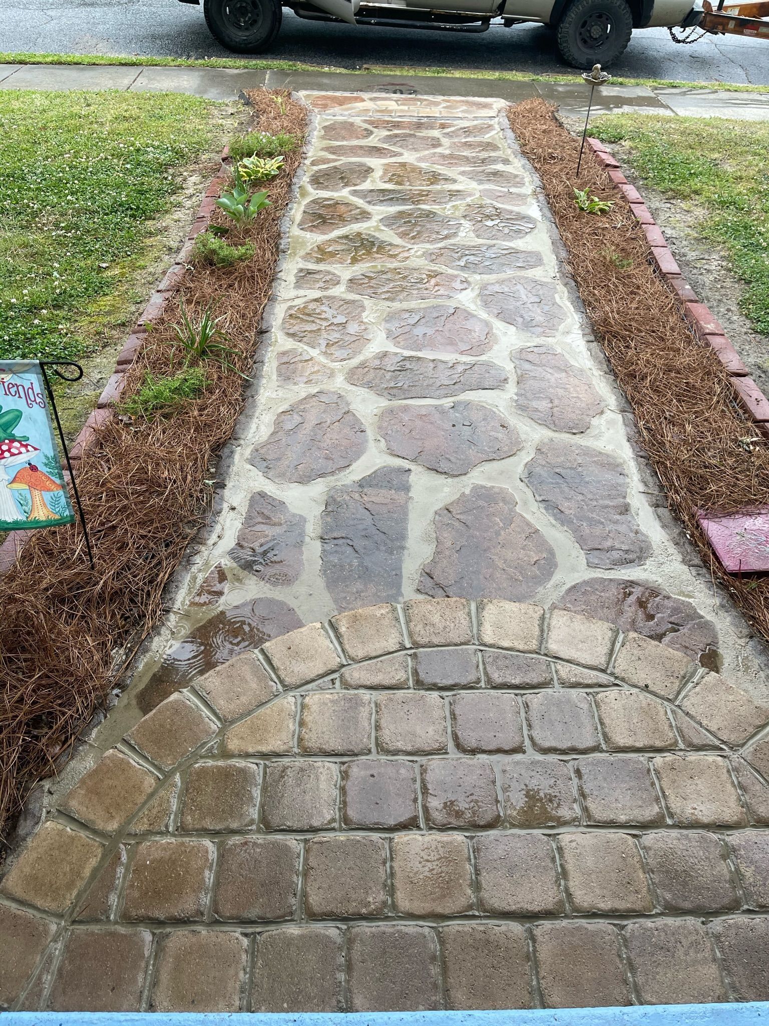 Stone walkway through a grassy yard leading to a street, bordered by red bricks and mulch.