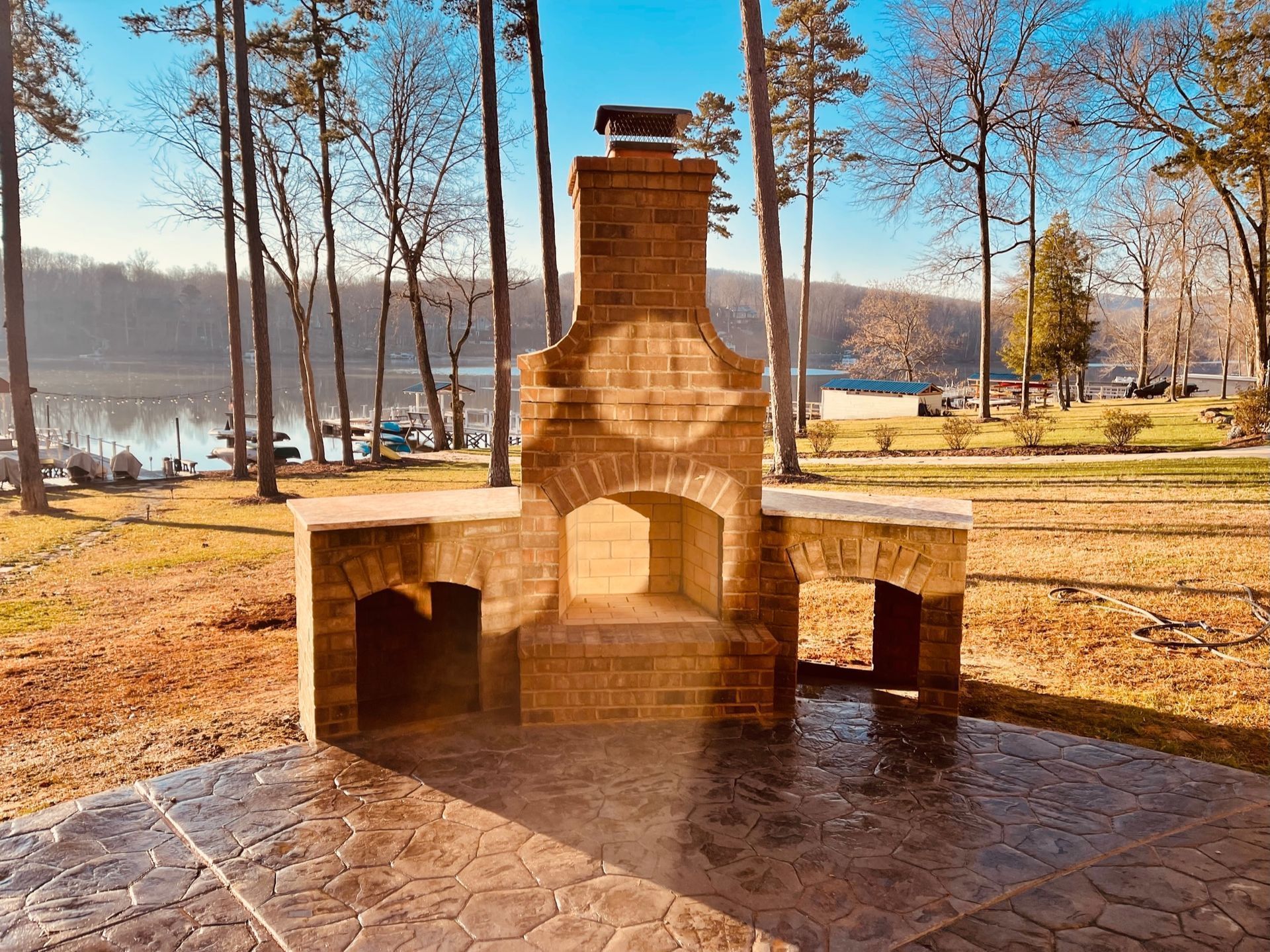 Stone outdoor fireplace and benches in a park by a lake, with leafless trees and sunlight.