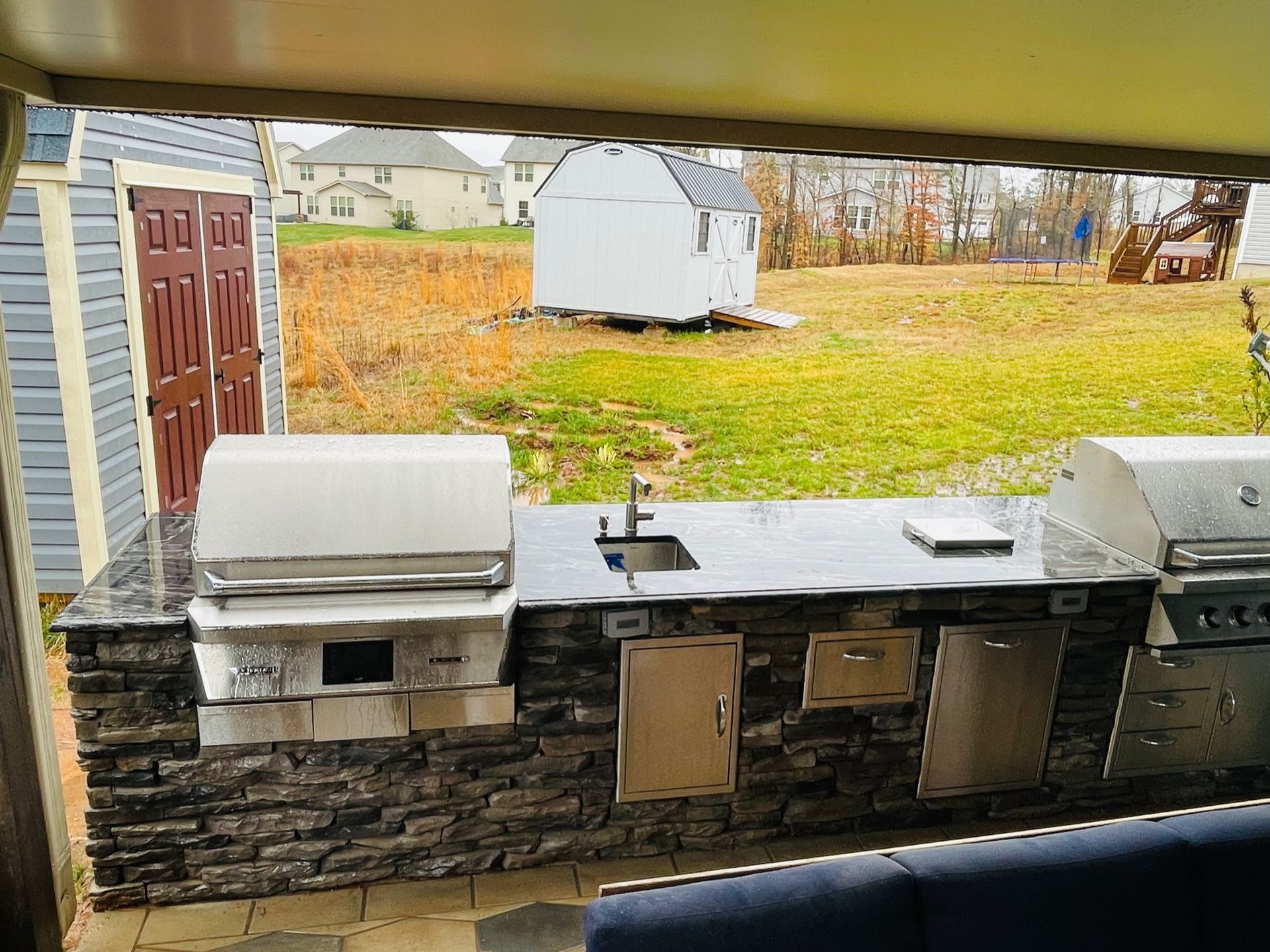 Outdoor patio with a stone counter, grills, and a sink overlooking a grassy yard and small white shed.