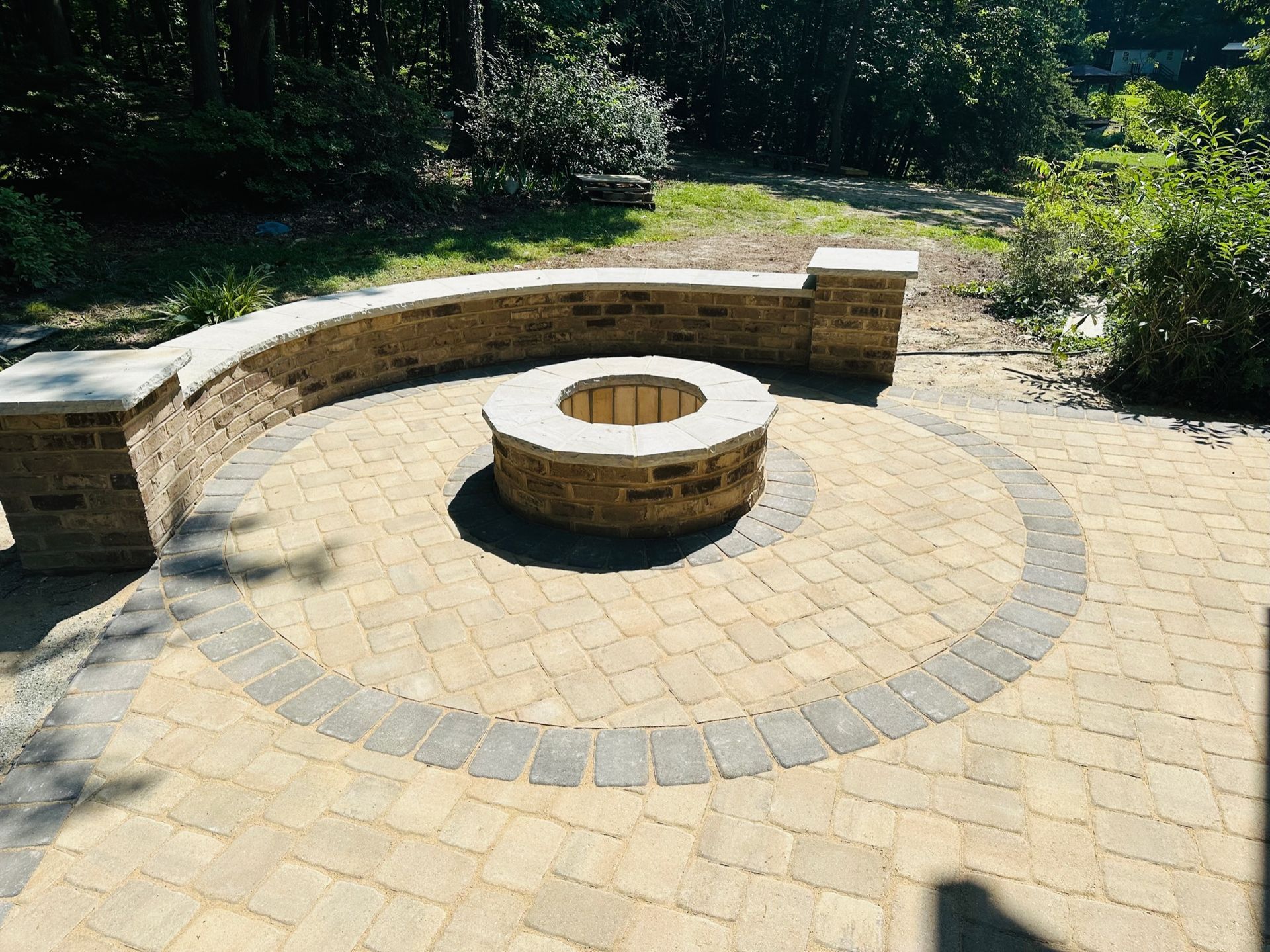 Curved stone bench around a circular fire pit in a sunlit brick patio, surrounded by greenery