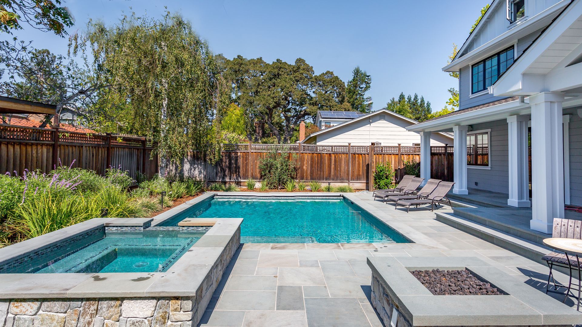Backyard with a turquoise pool, hot tub, stone patio, and a modern house with white columns