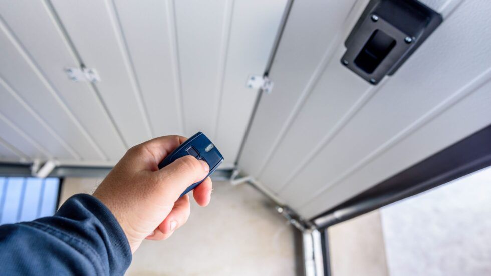 Hand pressing button on garage door opener remote; garage door and opener visible.