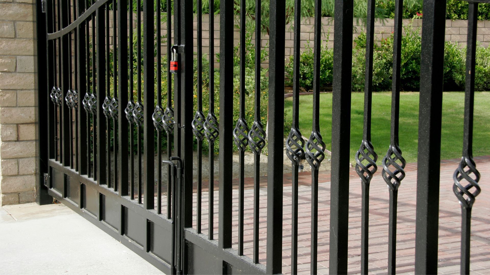 Black metal gate with decorative twists, locked, in front of a brick wall and grassy lawn.