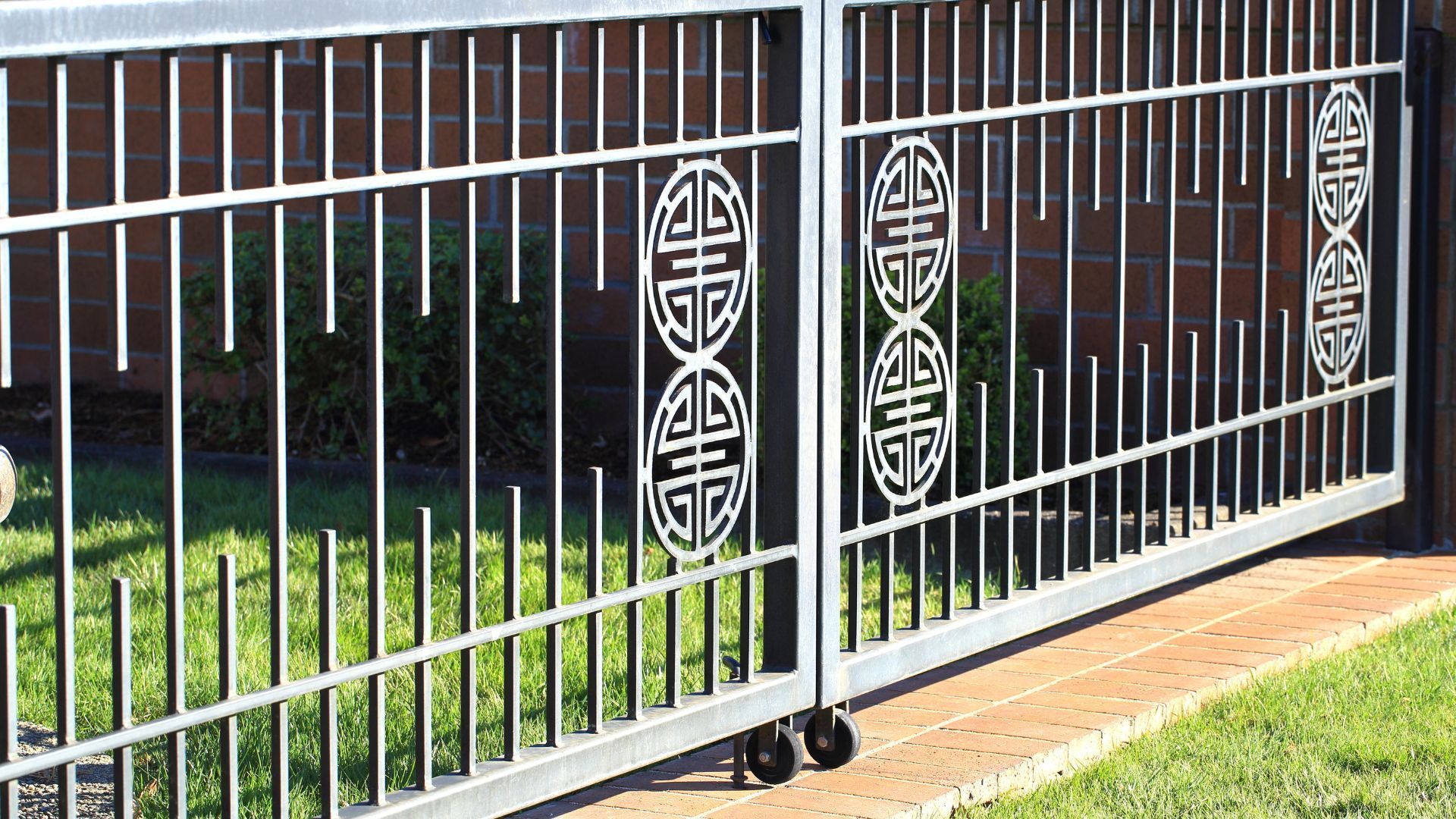 Sliding metal gate with vertical bars and decorative center designs, set against a brick wall and grass.