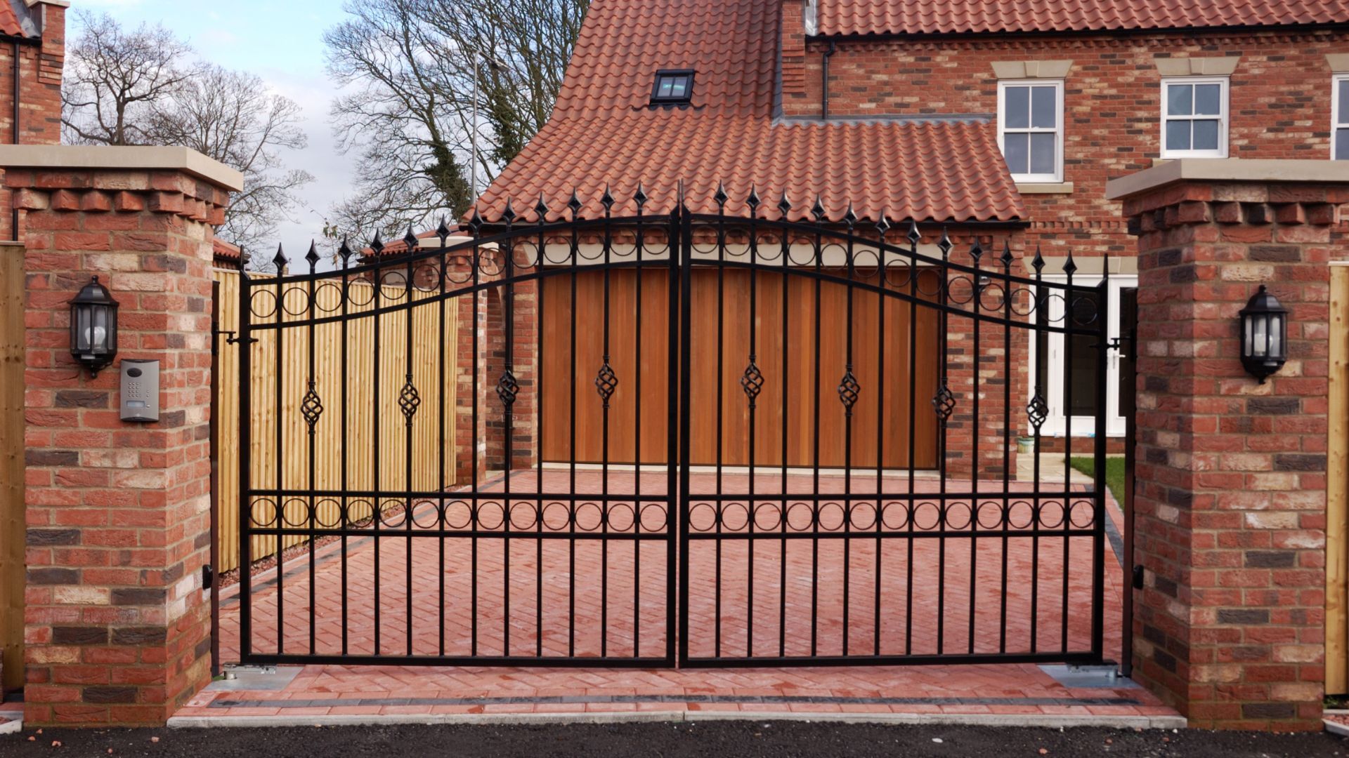 Black wrought iron gate with brick pillars in front of a house with red tiled roof. Black wrought iron gate with brick pillars in front of a house with red tiled roof.
