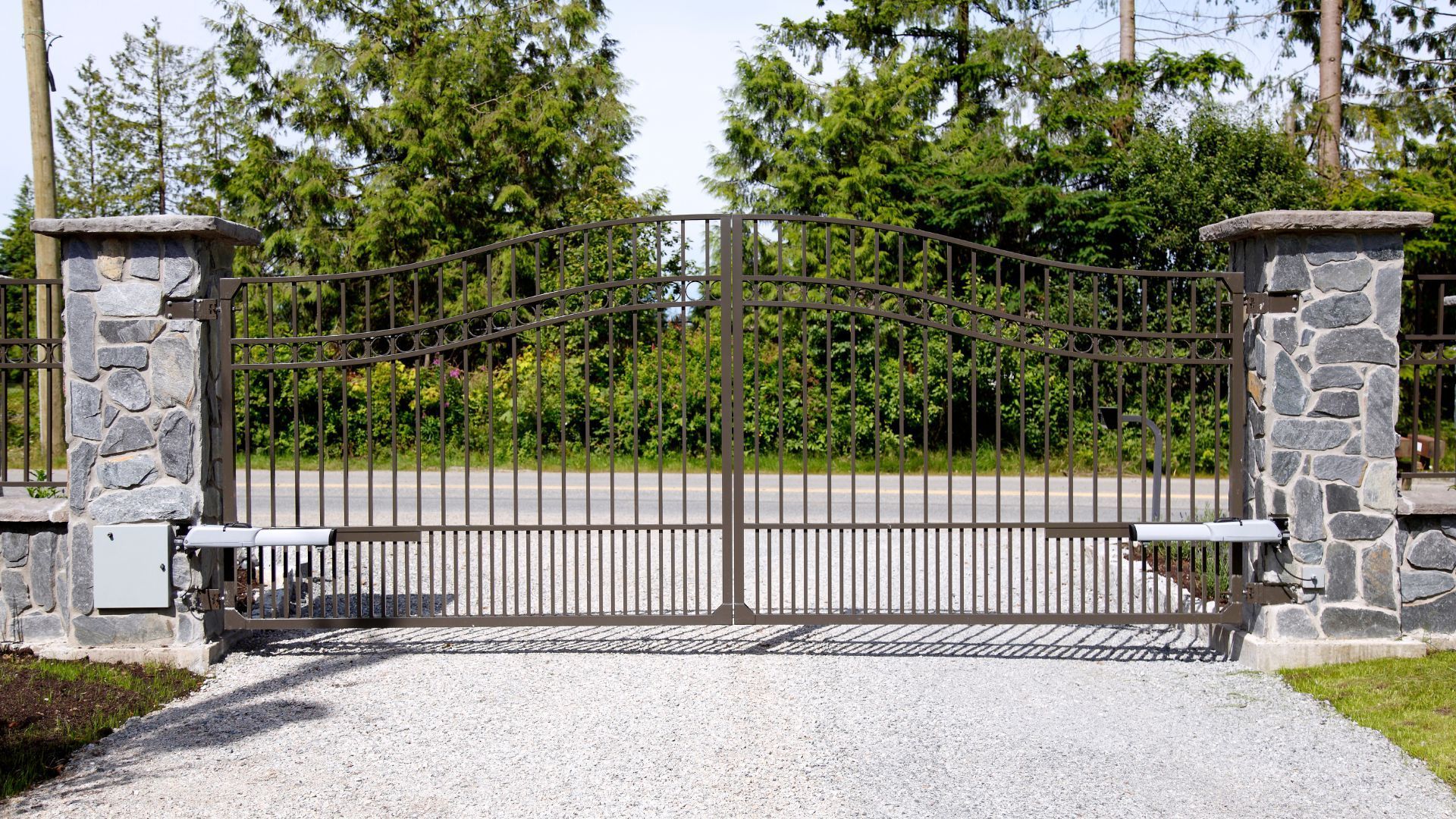 Iron gate with stone pillars, spanning a gravel driveway; trees in the background. Iron gate with stone pillars, spanning a gravel driveway; trees in the background.