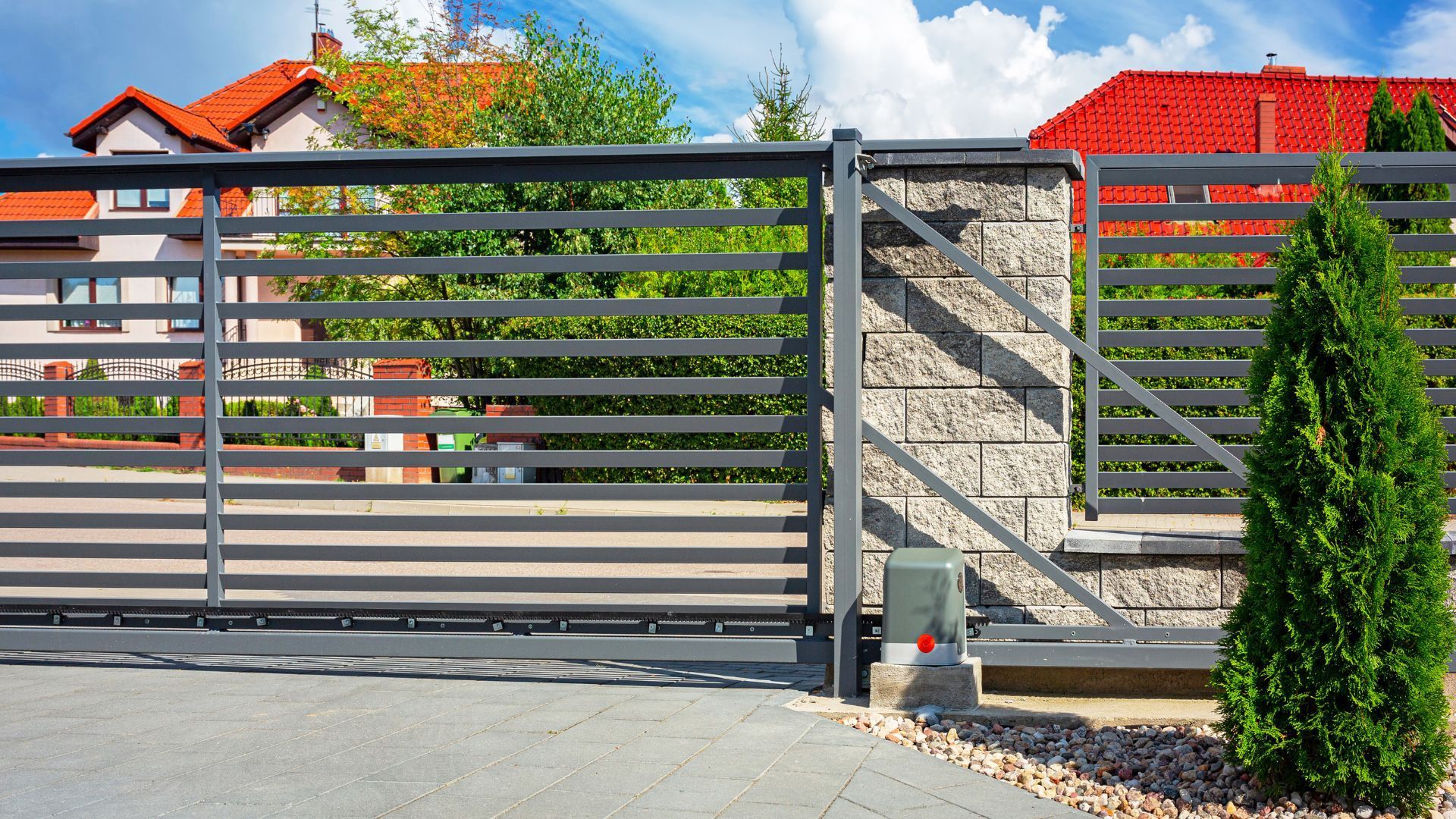 Gray sliding gate in front of a house with red roof, stone pillars, and green shrubs.