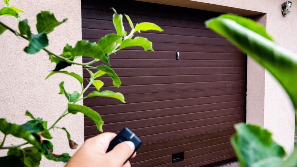 Hand using a remote to open a brown garage door. Green foliage in the foreground and on the side.