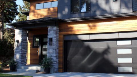 Modern home exterior with wooden siding, grey garage door, and stone columns.