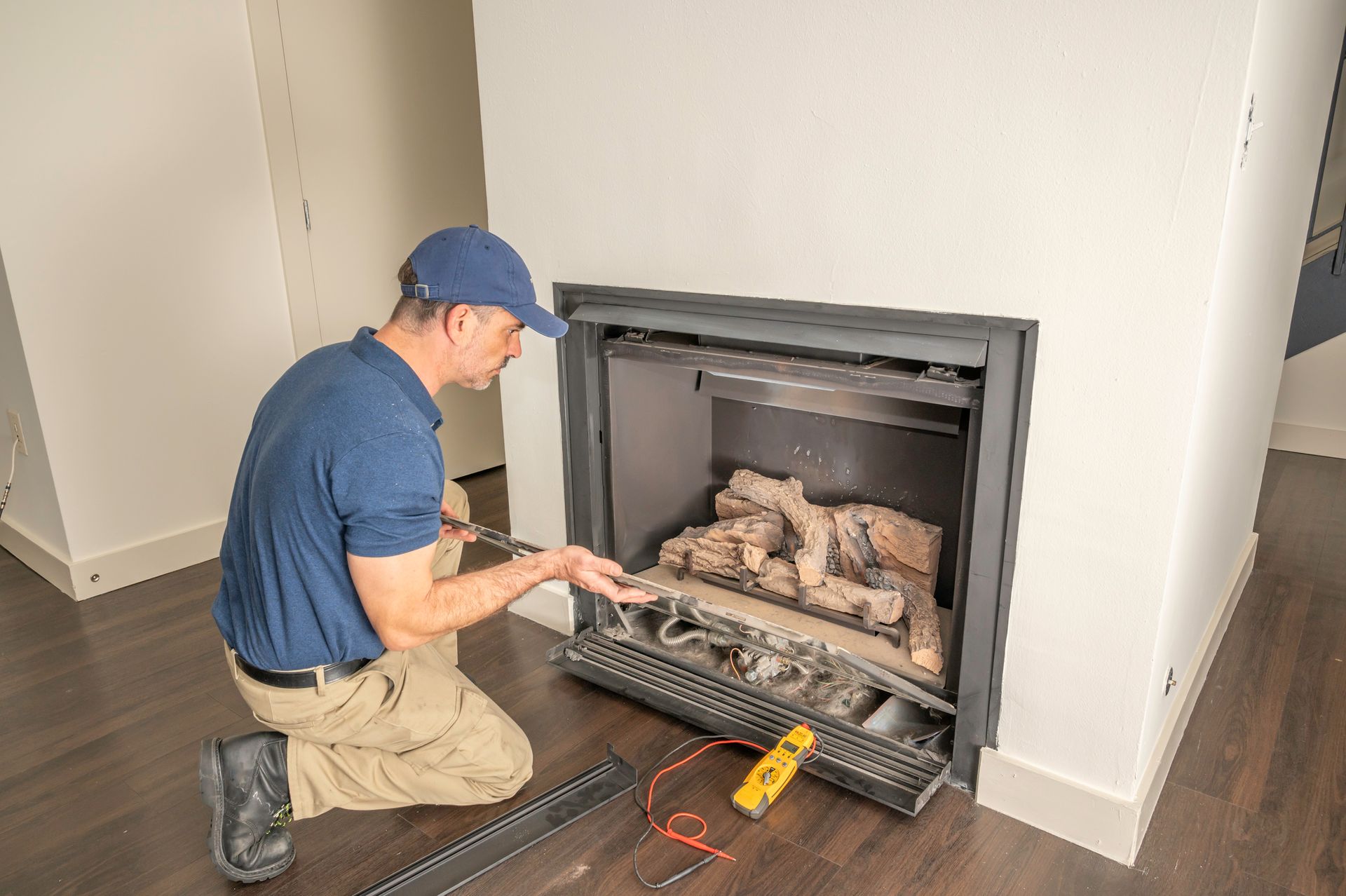 Man kneels, working on a fireplace. He wears a blue shirt and hat, using a tool.