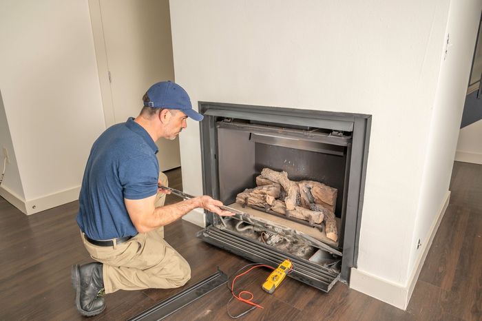 Man kneels, working on a fireplace. He wears a blue shirt and hat, using a tool.
