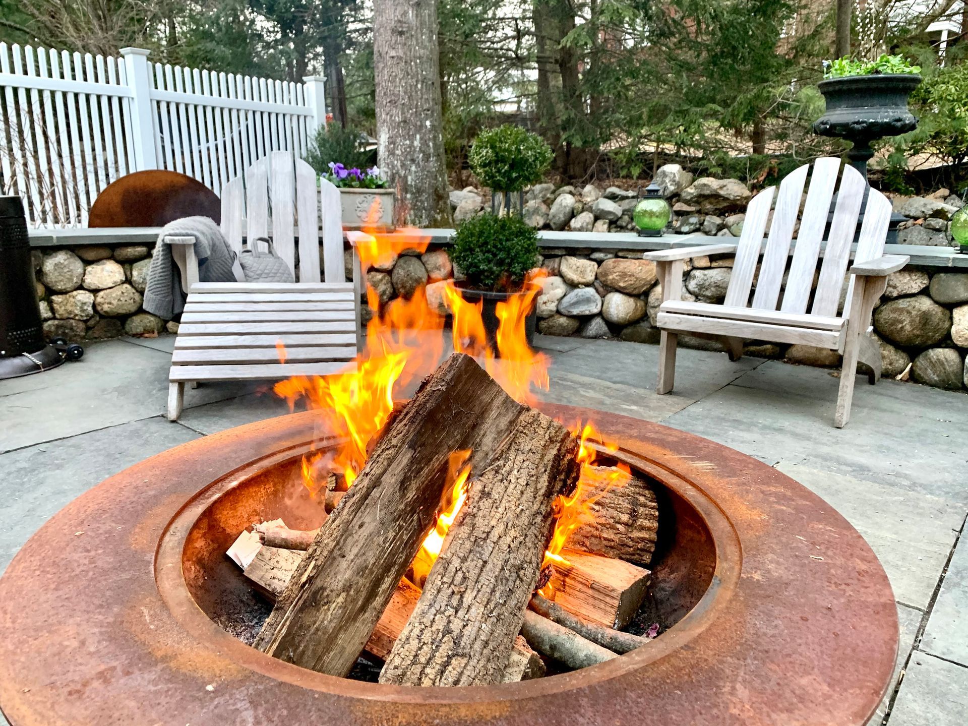 Fire pit with burning logs, two Adirondack chairs, stone wall, and white fence.