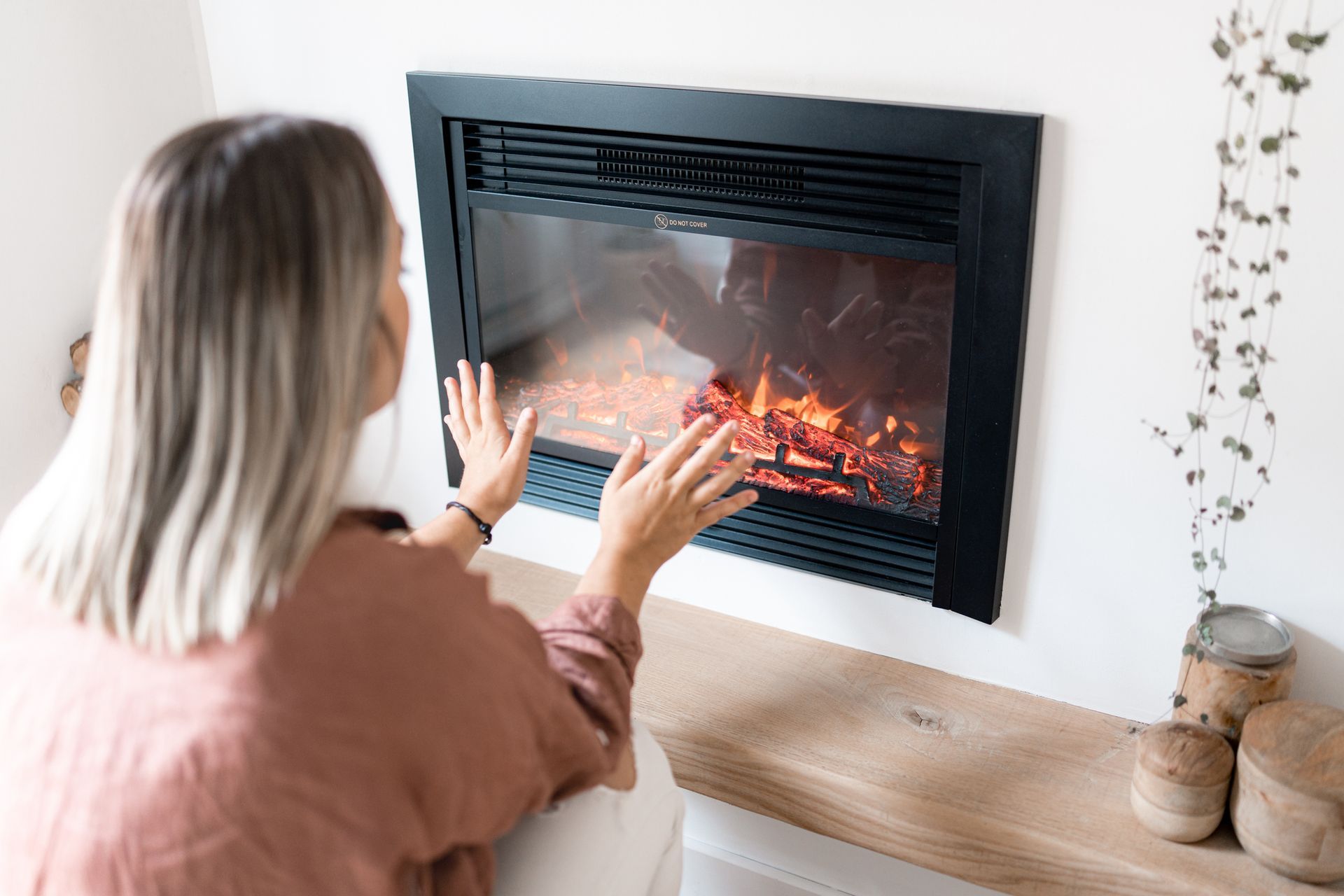 Woman warming hands near a black fireplace with a realistic flame effect in a white-walled room.