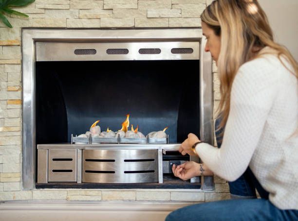 Woman adjusting a gas fireplace. She is near a brick wall with the fire lit.