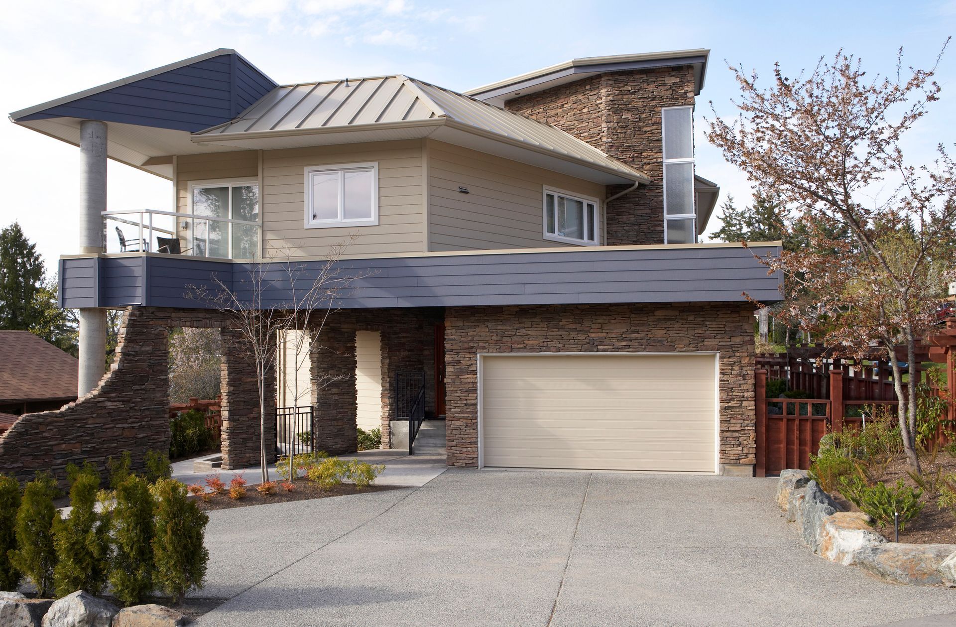 Modern two-story house with stone facade, tan siding, and a two-car garage. A driveway leads to the house, and trees are in view.