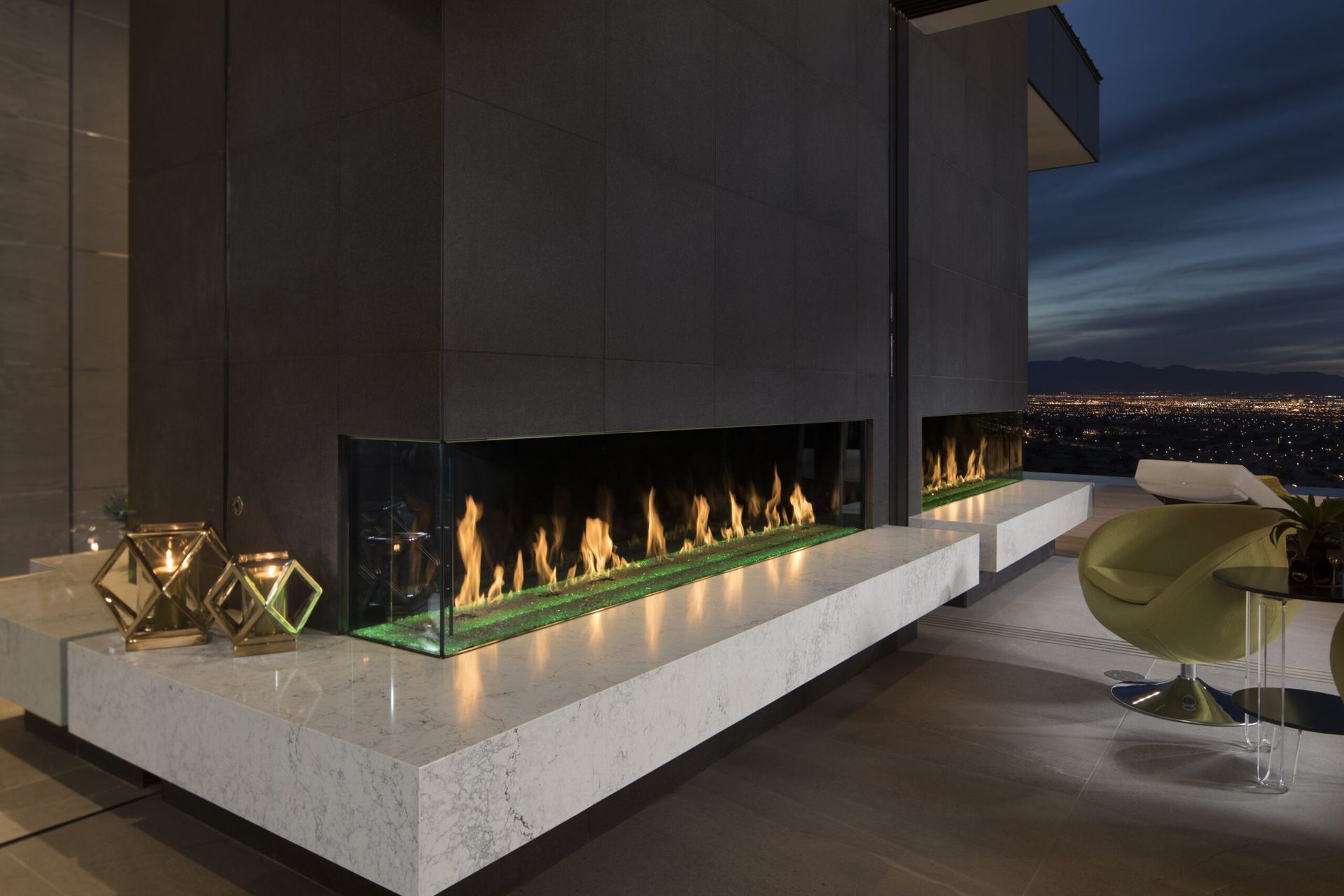 Modern fireplace with flames, built into a concrete ledge, overlooking city lights at dusk.