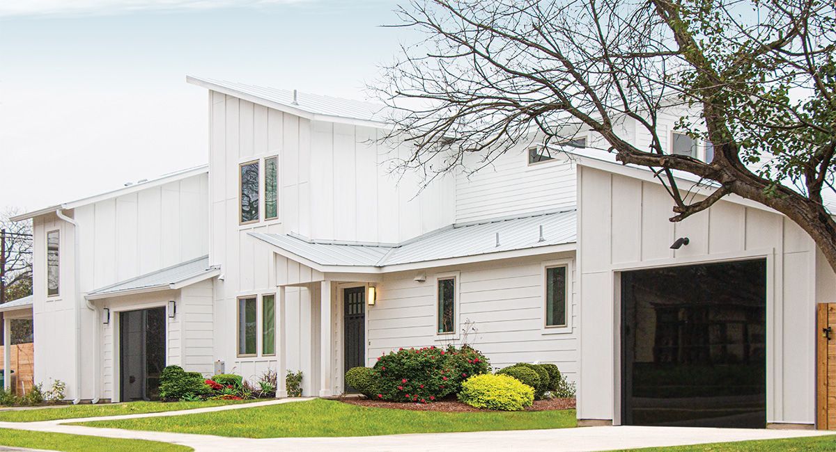 White modern house with a garage and angled roof. Green lawn, small shrubs, and tree visible.
