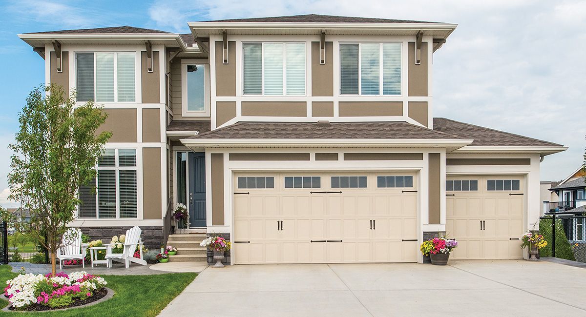 Two-story brown house with cream trim and garage doors, front yard with flower bed and chairs.