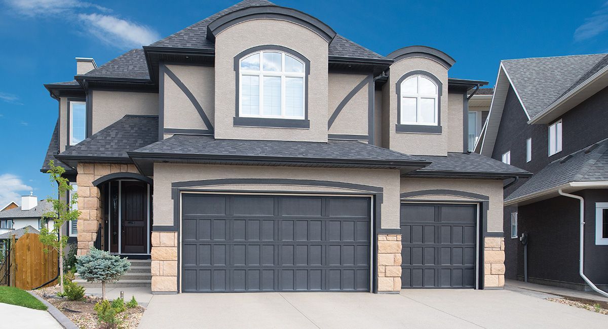 Two-story house with gray stucco exterior, dark gray garage doors, and tan stone accents.