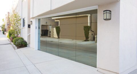 Glass garage door reflecting a nearby building and greenery on a concrete driveway.
