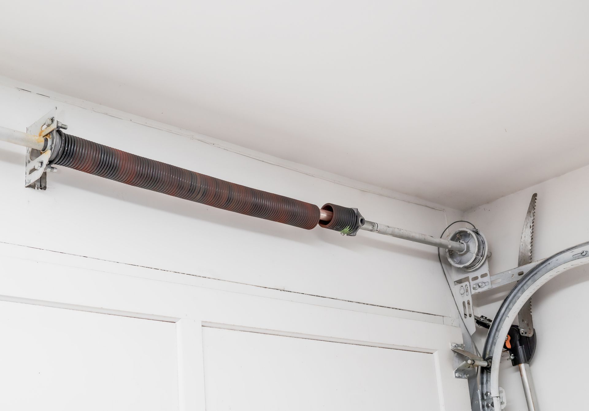 Garage door torsion spring, rusty, mounted horizontally above a white garage door.