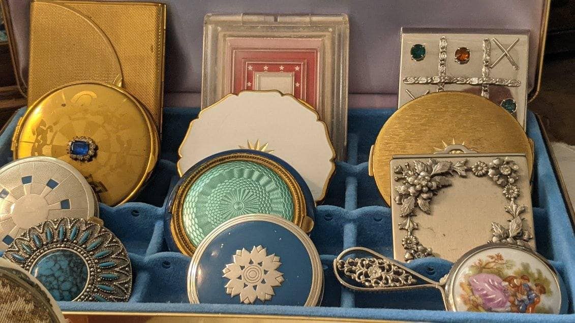 A collection of vintage compact mirrors in various shapes, colors, and ornate designs, arranged in a blue velvet tray.
