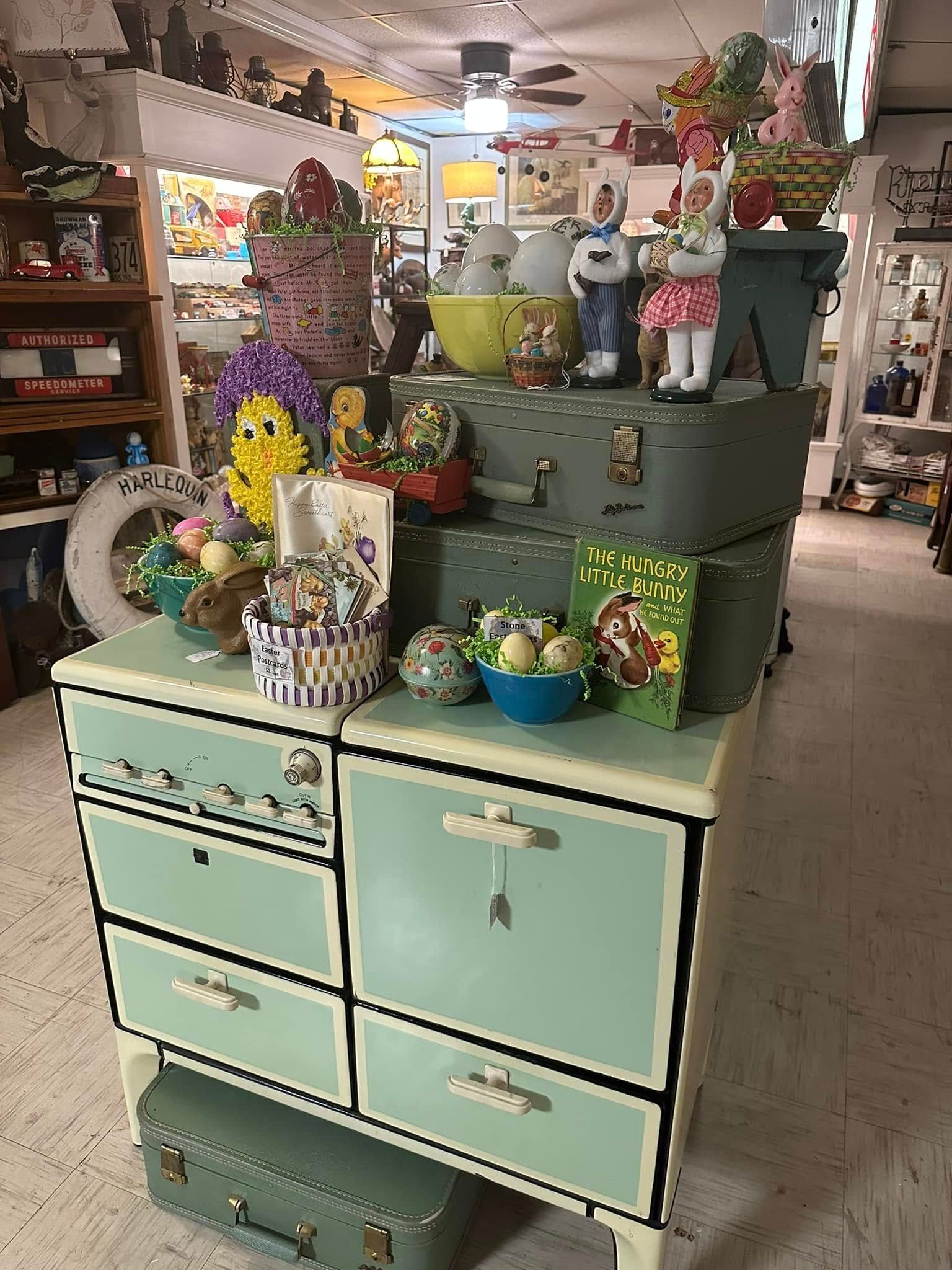 A vintage green cabinet topped with stacked suitcases, Easter-themed decor, and toy figures in an antique shop.