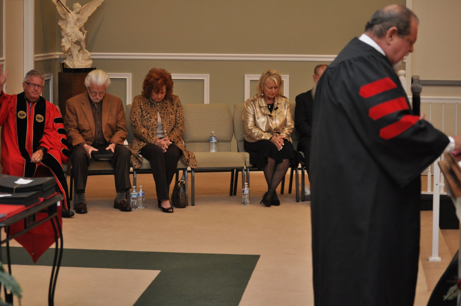 People in formal attire, some with heads bowed, at an event. A robed person is on the right.