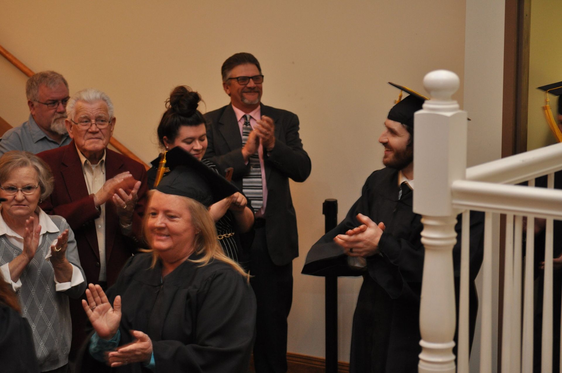 Graduates and audience clap.  A man in a cap and gown smiles on the stairs.  Indoors, festive setting.