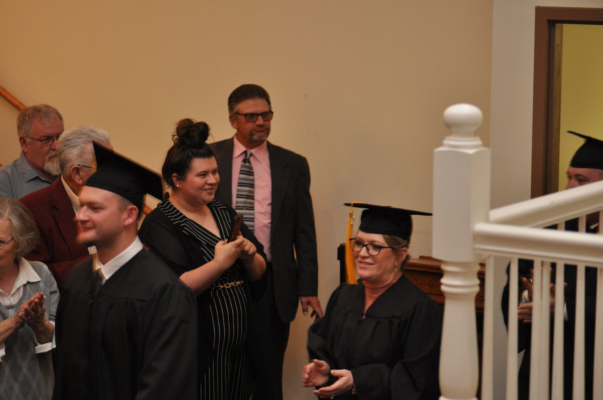 Graduates in black caps and gowns at a ceremony, smiling. A man in suit stands behind a woman. Interior with stairs.