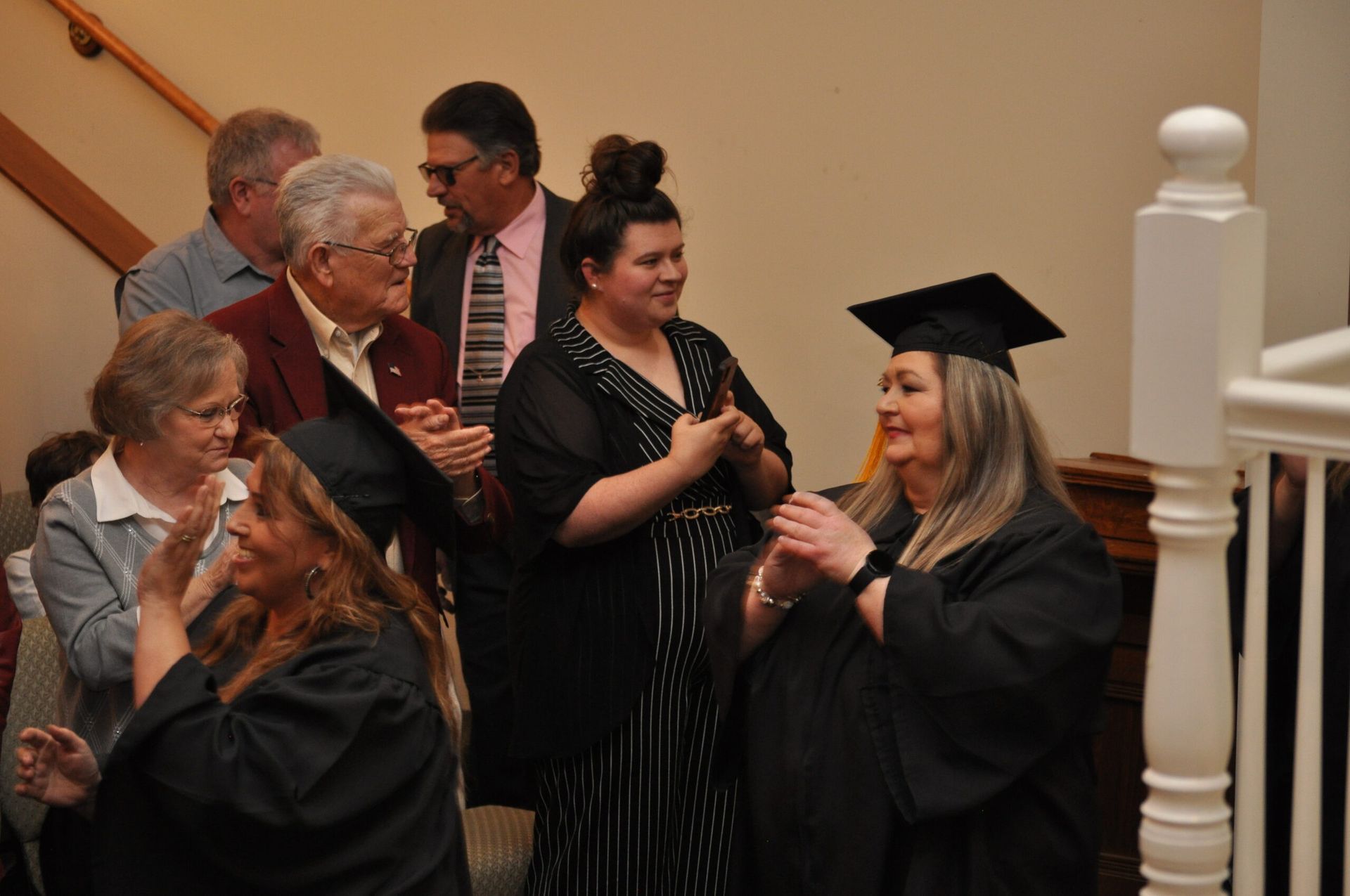 Graduates in black caps and gowns applaud, some with family, on a stairwell.