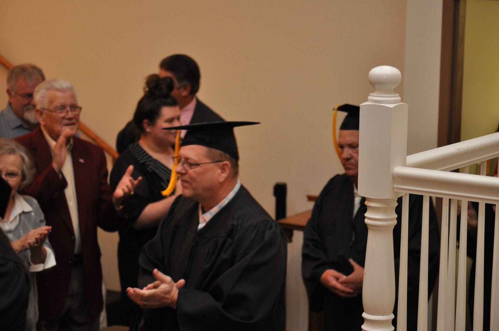 Graduates in caps and gowns descend a staircase, applauded by onlookers in a brightly lit hallway.