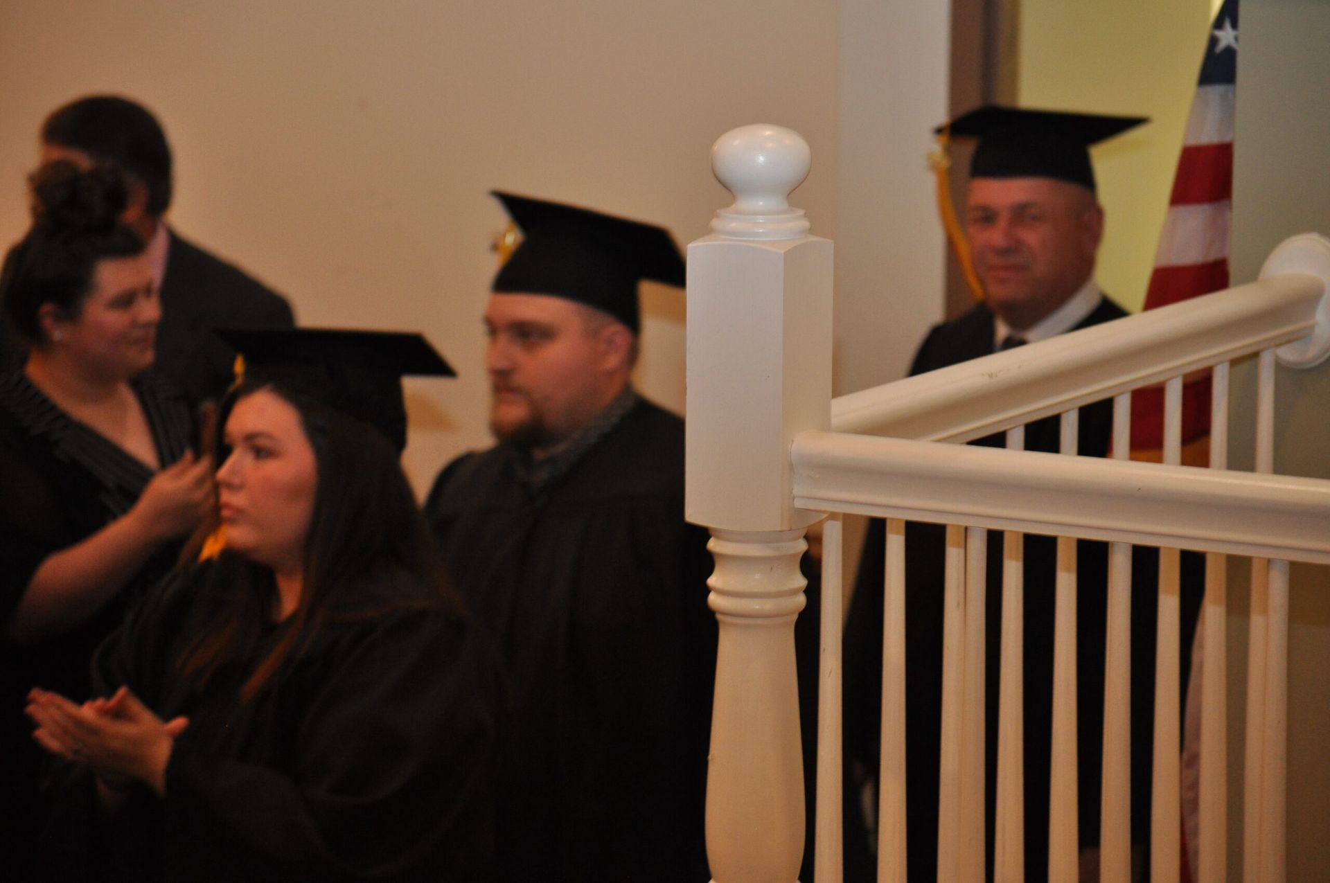 Graduates in black caps and gowns wait on stairs, near an American flag.