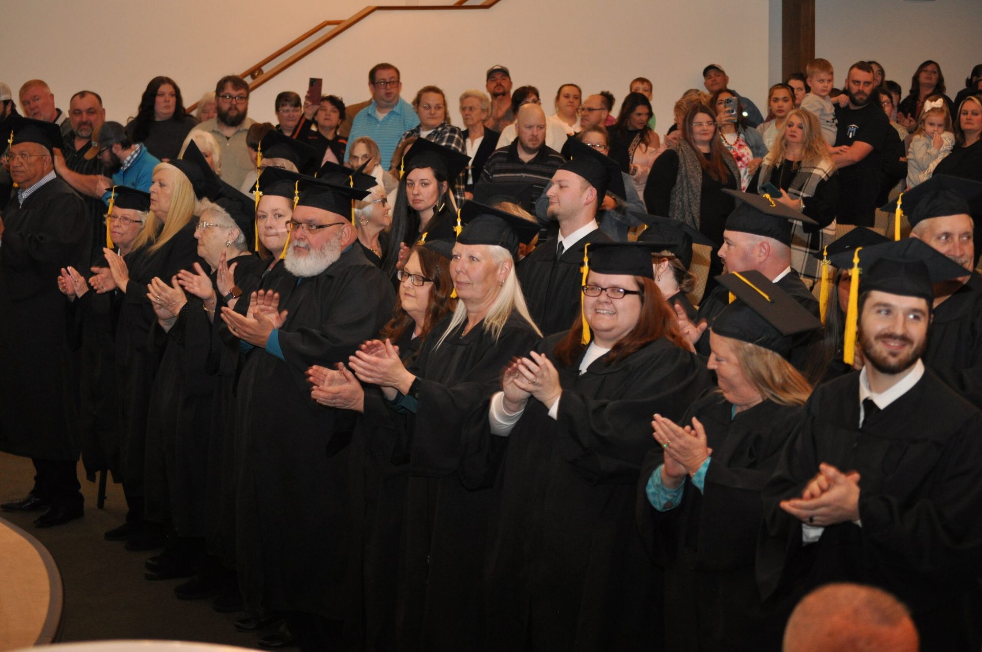 Graduates in black gowns and caps clapping, facing a crowd at a ceremony.
