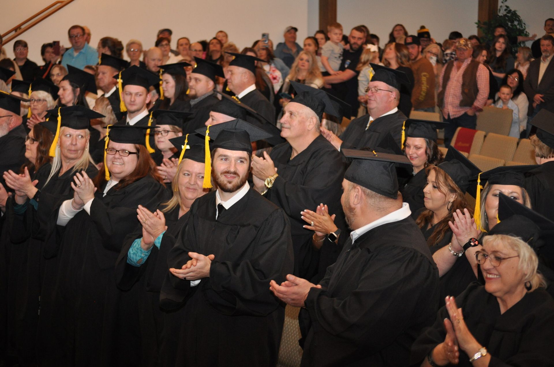 Graduates in black gowns and mortarboards applaud during a ceremony.