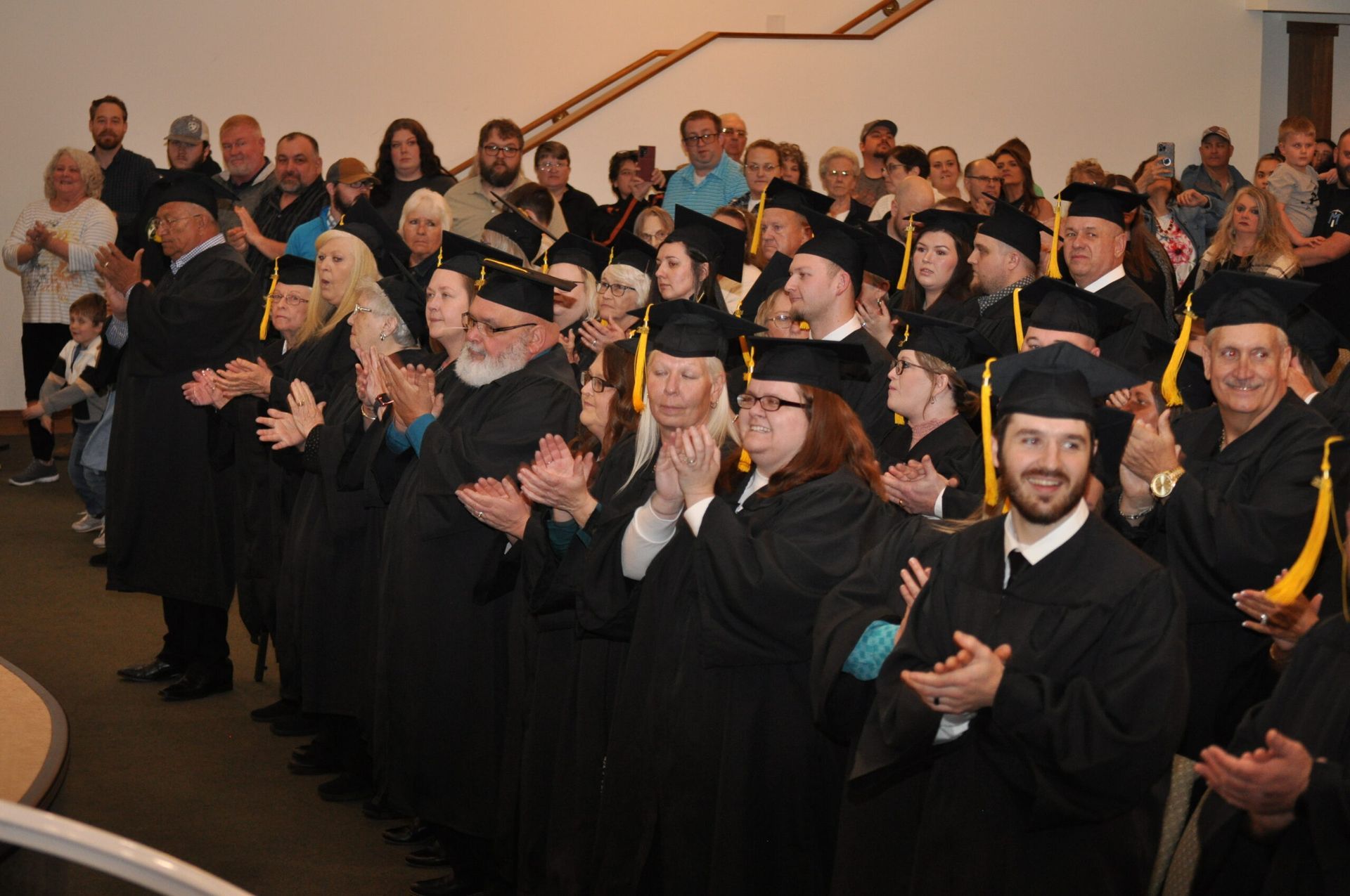 Graduation ceremony. Graduates in black gowns and caps clapping, audience in background.