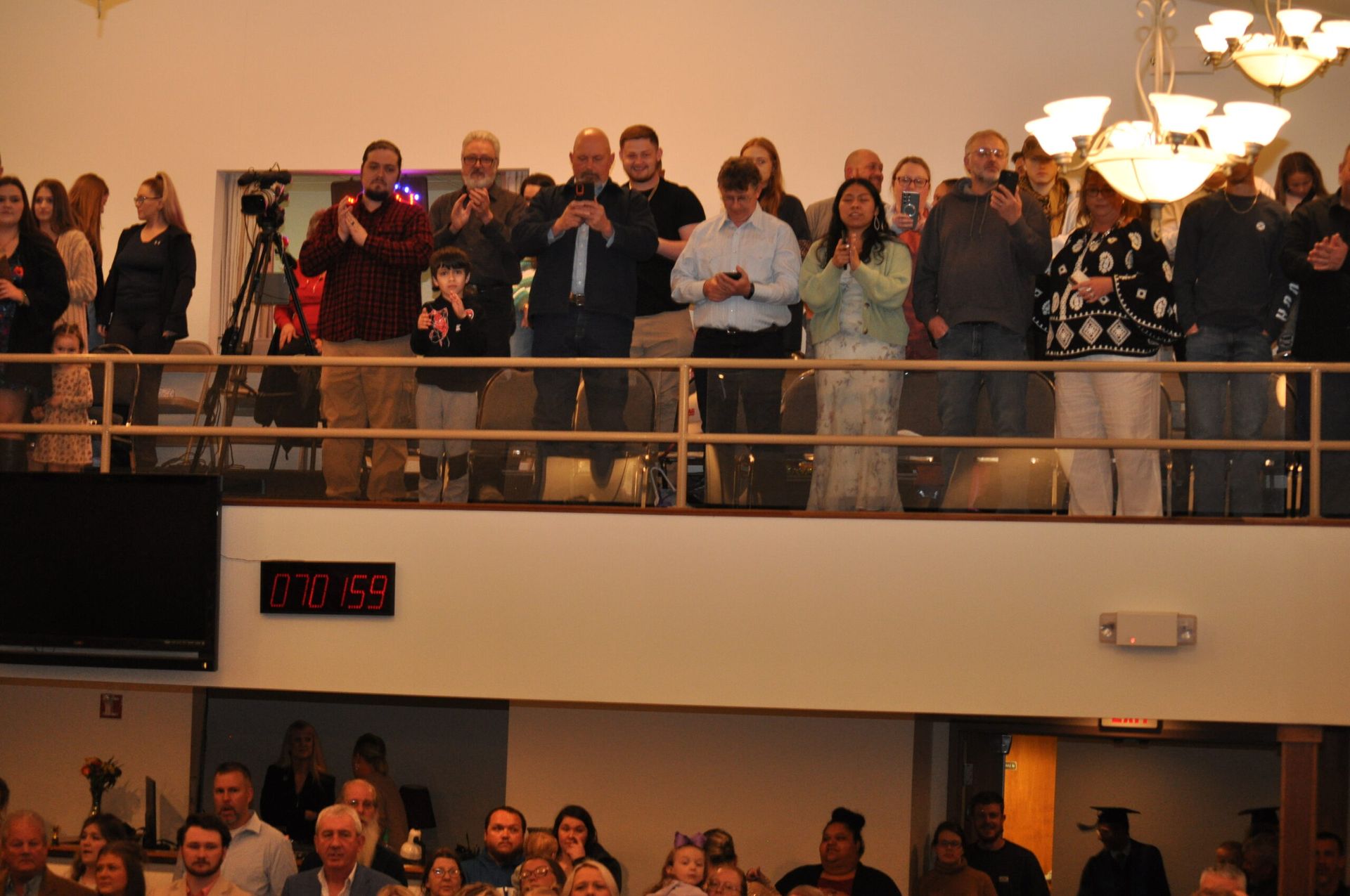 People standing on a balcony, clapping and looking down at a crowd in a well-lit room.